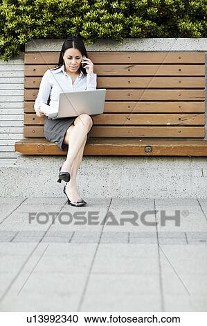 Stock Image - Young businesswoman sitting on bench using laptop. Fotosearch