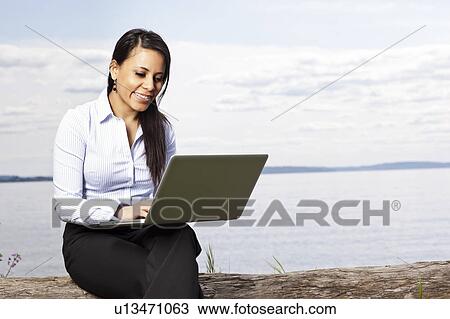 Stock Image - Young businesswoman using laptop on lakeshore. Fotosearch