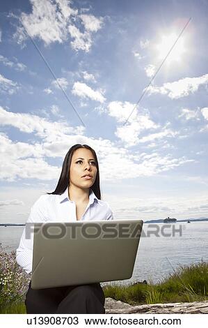 Stock Image - Young businesswoman using laptop on lakeshore. Fotosearch