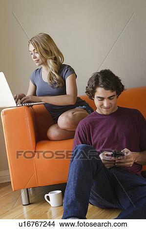 Picture - Young couple in living room, using phone and laptop. Fotosearch
