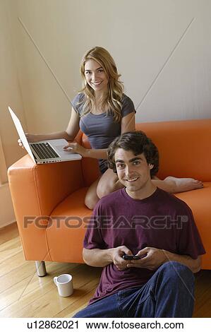Stock Image - Young couple in living room, using phone and laptop. Fotosearch