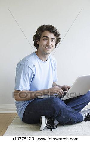 Stock Image - Young man sitting in room using laptop. Fotosearch