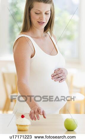 Stock Image - Young pregnant choosing between cupcake and apple. Fotosearch