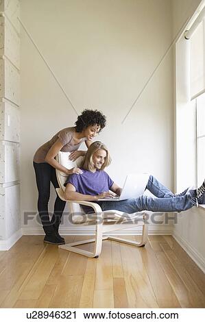 Stock Image - Young woman looking at man using laptop at home. Fotosearch