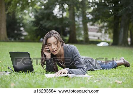 Young woman lying on grass using laptop and cell phone View Large Photo Image Picture - Young woman lying on grass using laptop and cell phone. Fotosearch