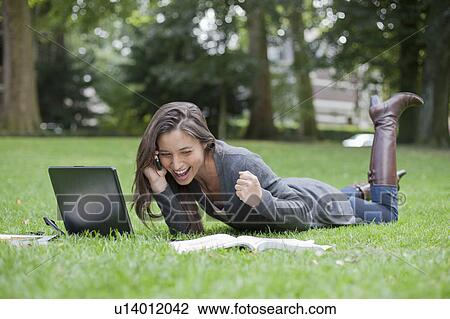 Stock Image - Young woman lying on grass using laptop and cell phone. Fotosearch