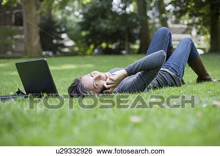 Young woman lying on grass using laptop and cell phone View Large Photo Image Stock Photograph - Young woman lying on grass using laptop and cell phone. Fotosearch