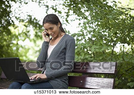 Young woman sitting on park bench using laptop and cell phone View Large Photo Image Stock Image - Young woman sitting on park bench using laptop and cell phone. Fotosearch