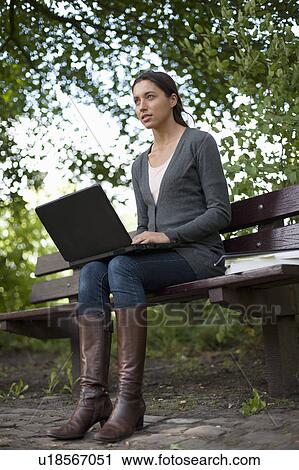 Young woman sitting on park bench using laptop View Large Photo Image Stock Image - Young woman sitting on park bench using laptop. Fotosearch