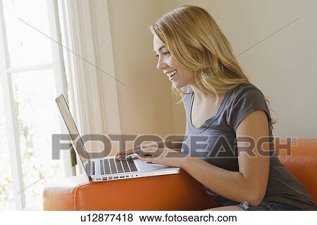 Stock Photo - Young woman sitting on sofa using laptop. Fotosearch