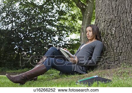Stock Image - Young woman sitting under tree reading books. Fotosearch