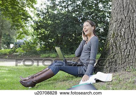 Stock Photo - Young woman sitting under tree using laptop and cell phone. Fotosearch