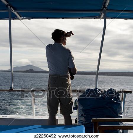 Stock Image - Man standing on a boat looking at a view, Santiago Island, Galapagos Islands, Ecuador. Fotosearch