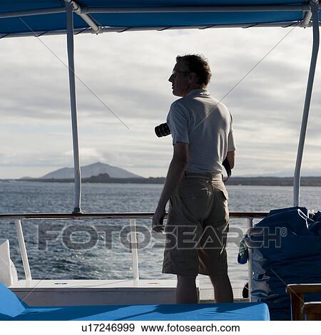 Stock Photo - Man standing on a boat looking at a view, Santiago Island, Galapagos Islands, Ecuador. Fotosearch