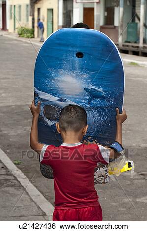 Stock Photograph - Rear view of a boy carrying a surfboard, Puerto Baquerizo Moreno, San Cristobal Island, Galapagos Islands, Ecuador. Fotosearch