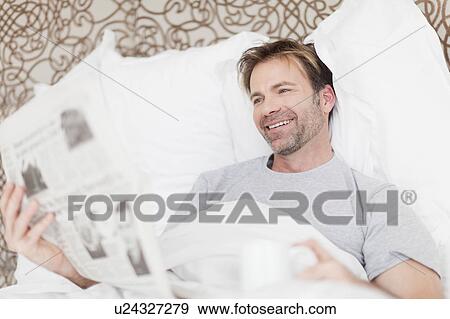 Smiling man reading newspaper in bed View Large Photo Image Stock Photo - Smiling man reading newspaper in bed. Fotosearch