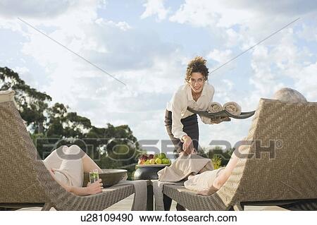 Stock Image - Waitress serving couple relaxing by pool. Fotosearch