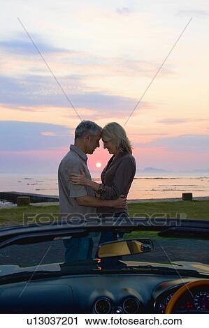 Stock Image - Couple hugging by convertible at sunset. Fotosearch