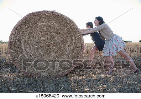 Stock Image - Girls pushing hay bale in field. Fotosearch