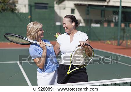 Stock Image - Older women hugging on tennis court. Fotosearch