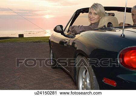 Stock Image - Woman sitting in convertible at sunset. Fotosearch