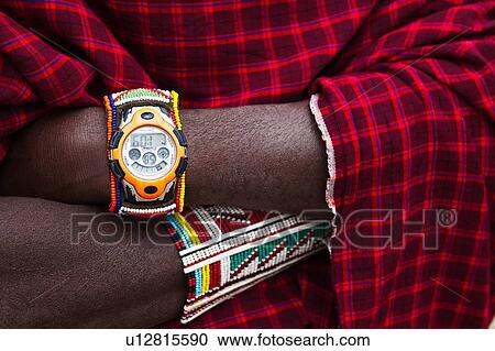 Stock Image - Close up detail of Masai man's arms folded, with beaded jewellery and beaded watch strap, background of traditional red shuka, Amboseli, Kenya, East Africa. Fotosearch