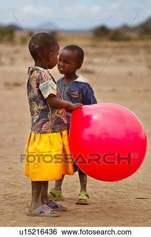 Stock Photograph - Close up detail of Masai two children at outdoor school one little girl at front holding a very large red balloon, and the one wishing it was hers? ÇªAmboseli, Kenya, East Africa. Fotosearch