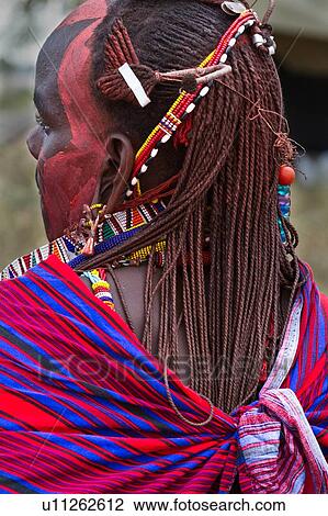 Stock Image - Close up side back view of young Masai warrior (moran), with long braided hair, wearing traditional red shuka and beaded jewellery, with red facial paint, Amboseli, Kenya, East Africa. Fotosearch