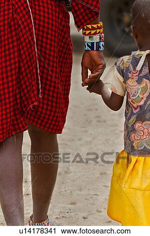 Stock Image - Father and child: detail of Masai man in traditional red shuka holding hand of his small daughter in bright yellow skirt, walking together, back view, Amboseli, Kenya, East Africa. Fotosearch