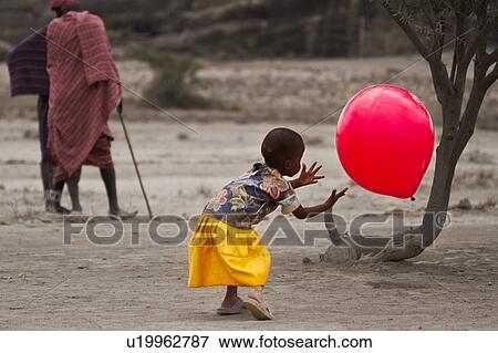 Stock Photo - Little girl playing outdoors with large red balloon, framed by tree trunk, while two Masai men talk in background, both dressed in traditional shukas, and one leaning on stick, Amboseli, Kenya, East Africa. Fotosearch