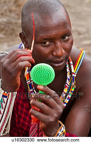 Stock Image - Masai applying paints for celebrations: Masai warrior dressed in traditional red shuka and much beaded jewellery sits applying red paint to his face with reed, using green plastic mirror Amboseli, Kenya, East Africa. Fotosearch