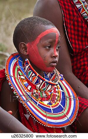 Stock Image - Masai applying paints for celebrations: child dressed in colourful clothing and much beaded jewellery sits after red paint has been applied to her face, Amboseli, Kenya, East Africa. Fotosearch