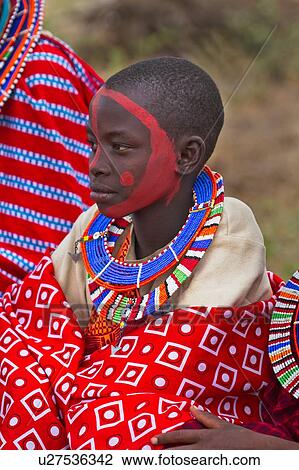 Stock Image - Masai applying paints for celebrations: child dressed in colourful clothing and much beaded jewellery sits after red paint has been applied to her face, Amboseli, Kenya, East Africa. Fotosearch