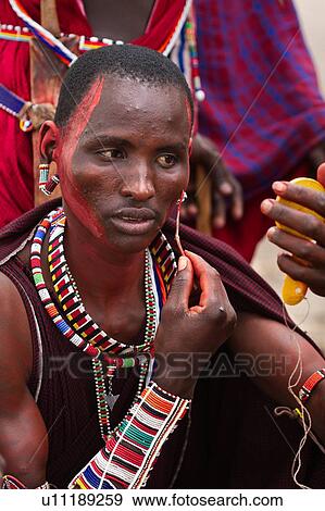 Masai applying paints for celebrations: Masai warrior dressed in traditional red shuka and much beaded jewellery sits applying red paint to his face with reed, using yellow plastic mirror Amboseli, Kenya, East Africa View Large Photo Image Stock Photo - Masai applying paints for celebrations: Masai warrior dressed in traditional red shuka and much beaded jewellery sits applying red paint to his face with reed, using yellow plastic mirror Amboseli, Kenya, East Africa. Fotosearch