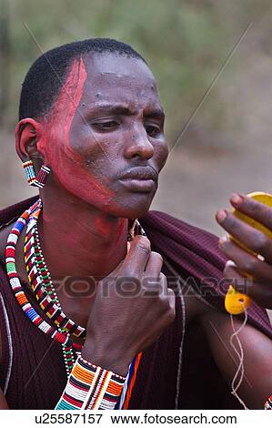 Stock Photo - Masai applying paints for celebrations: Masai warrior dressed in traditional red shuka and much beaded jewellery sits applying red paint to his face with reed, using yellow plastic mirror Amboseli, Kenya, East Africa. Fotosearch