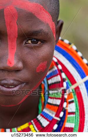 Stock Photograph - Masai applying paints for celebrations: young woman with much beaded jewellery sits after red paint has been applied to her face, close up detail of face, Amboseli, Kenya, East Africa. Fotosearch