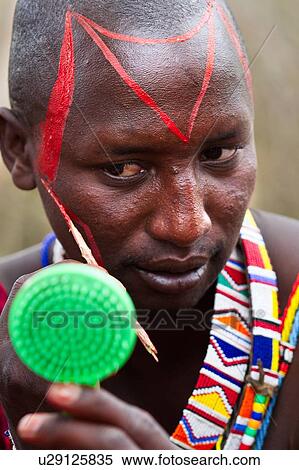 Masai applying paints for celebrations: Masai warrior dressed in traditional red shuka and much beaded jewellery sits applying red paint to his face with reed, using green plastic mirror Amboseli, Kenya, East Africa View Large Photo Image Stock Photography - Masai applying paints for celebrations: Masai warrior dressed in traditional red shuka and much beaded jewellery sits applying red paint to his face with reed, using green plastic mirror Amboseli, Kenya, East Africa. Fotosearch