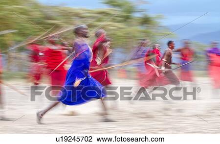 Stock Image - Masai Olympics: group of Masai warriors, all dressed in traditional red and blue shukas, run with spears and sticks in spear throwing competition, motion blur effect, Amboseli, Kenya, East Africa. Fotosearch