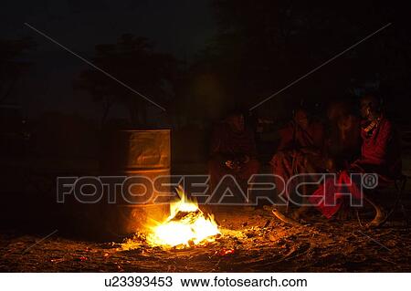 Stock Image - Masai safari camp: Masai sit around camp fire, dressed in traditional red shukas, jeeps in background, tall trees beyond, Amboseli, Kenya, East Africa. Fotosearch