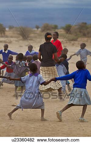 Stock Image - Masai school out on the plains: a group of children, mostly dressed in blue sweaters, and blue check clothing dance in a circle around their two teachers, dressed in red, and in black, Amboseli, Kenya, East Africa. Fotosearch