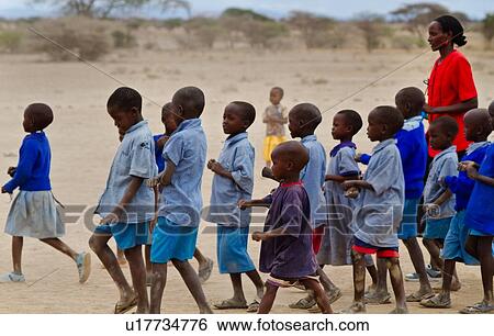 Stock Photograph - Masai school out on the plains: as seen from the side, a group of children, mostly dressed in blue sweaters, and blue check, march with their teacher, dressed in red, Amboseli, Kenya, East Africa. Fotosearch