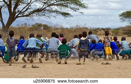 Stock Photography - Masai school out on the plains: as seen from behind, a group of children, mostly dressed in blue sweaters, and blue check clothing, pretend to hop like frogs, Amboseli, Kenya, East Africa. Fotosearch