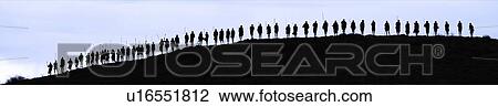 Stock Image - Masai silhouettes: over fifty Masai warriors stand at top of hill in early evening, holding traditional sticks, outlined against bright sky, Amboseli, Kenya, East Africa. Fotosearch