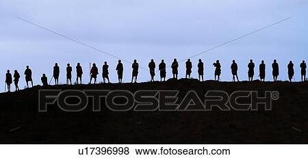 Stock Photo - Masai silhouettes: over twenty Masai warriors stand at top of hill in early evening, holding traditional sticks, outlined against bright sky, Amboseli, Kenya, East Africa. Fotosearch