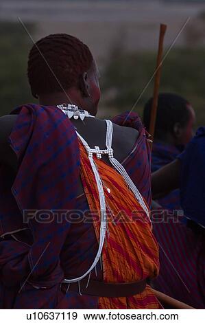 Masai warrior at dusk: close up back view as he sits looking to his right, wearing traditional blue and red shuka, orange-red tunic, and white beads, face painted with red, Amboseli, Kenya, East Africa View Large Photo Image Stock Photo - Masai warrior at dusk: close up back view as he sits looking to his right, wearing traditional blue and red shuka, orange-red tunic, and white beads, face painted with red, Amboseli, Kenya, East Africa. Fotosearch