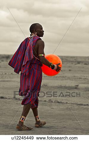 Masai warrior, dressed in traditional red and blue shuka walking away, diagonally, holding an orange balloon: desaturated image, Amboseli, Kenya, East Africa View Large Photo Image Stock Photo - Masai warrior, dressed in traditional red and blue shuka walking away, diagonally, holding an orange balloon: desaturated image, Amboseli, Kenya, East Africa. Fotosearch