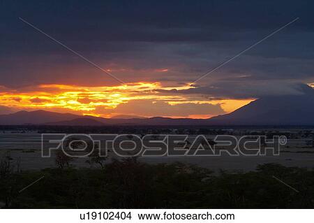 Sunset at Amboseli: dramatic red orange yellow against stormy grey clouds, over distant mountain, sandy plains and trees in foreground, Amboseli, Kenya, East Africa View Large Photo Image Picture - Sunset at Amboseli: dramatic red orange yellow against stormy grey clouds, over distant mountain, sandy plains and trees in foreground, Amboseli, Kenya, East Africa. Fotosearch
