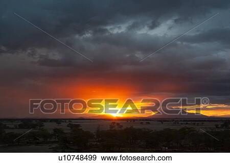 Stock Photo - Sunset at Amboseli: dramatic red orange yellow against stormy grey clouds, over distant mountain, sandy plains and trees in foreground, Amboseli, Kenya, East Africa. Fotosearch
