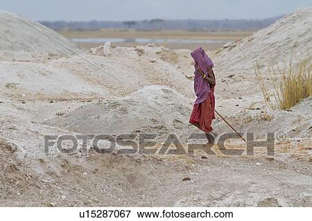 Stock Photo - Young Masai man with head covered by red and blue shuka stands in dusty dunes of Amboseli lake bed, framed by slopes, Amboseli, Kenya, East Africa. Fotosearch