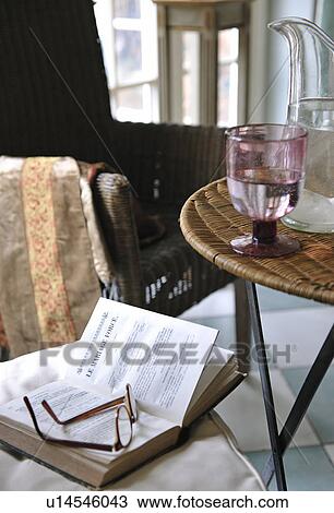 Stock Image - Book and eyeglasses on chair next to table with glass of water. Fotosearch